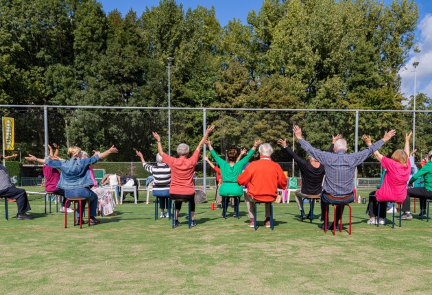 Ouderen volgen bij SKVR een dansles Zitdansen op een voetbalveld.