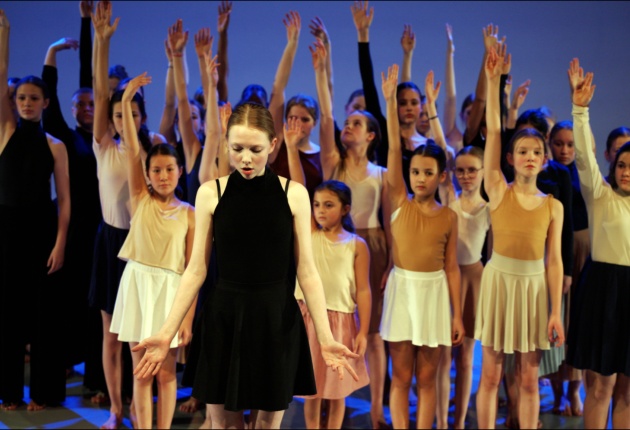 Groep jonge dansers op een podium, met één danser in het zwart vooraan en de rest met een arm omhoog. Ze dragen aardetinten en worden belicht door blauw licht.