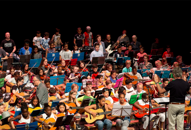 Een groot jeugdorkest speelt op een podium onder leiding van een dirigent, met diverse instrumenten zoals gitaren, violen en fluiten.